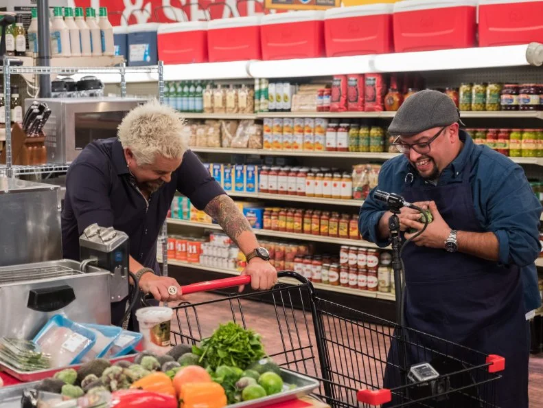 Host Guy Fieri laughs as Champion Carl Ruiz piles items into his cart during the West Coast - Single Aisle Showdown, as seen on Food Network's Guy's Grocery Games, Season 12.