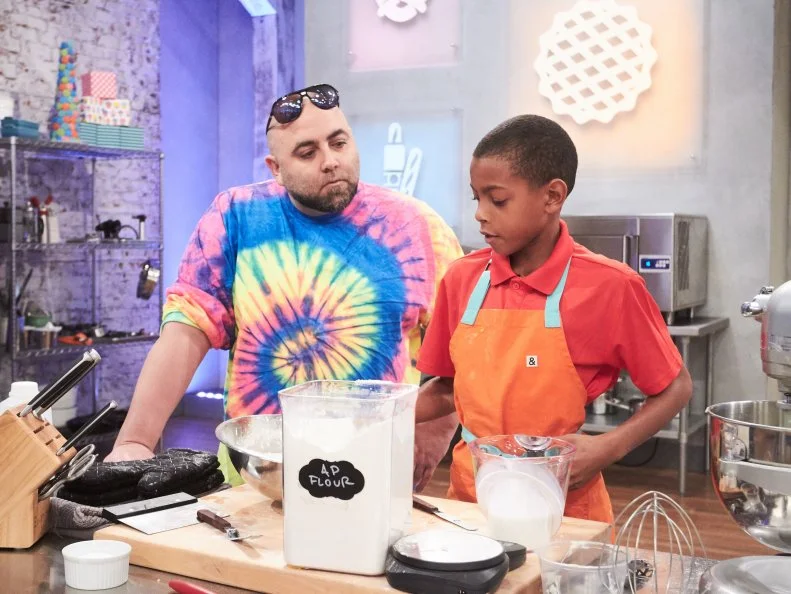 Host Duff Goldman watches contestant Reese Smith preparing his Main Heat dish, Tie Die Yellow and White Cake with American Buttercream and Vanilla Blackberry Swirl Ice Cream, as seen on Food Network's Kids Baking Championship, Season 3.