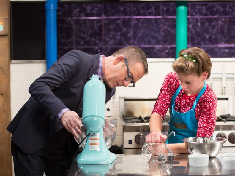 Host Ted Allen (L) helps out Junior contestants Nicole Bidun as she cooks Molton chocolate lave cake with a candied bacon and toasted brioche bun crumble and caramelized Sharon fruit during the dessert round, as seen on Food Network's Chopped Junior, Season 6.