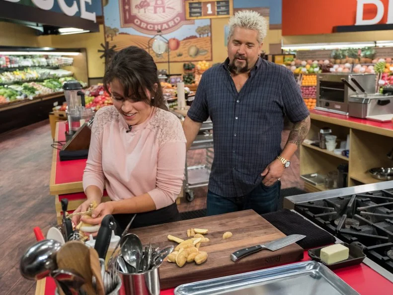 Host Guy Fieri checks in on Contestant Kate Newman as she prepares potatoes, as seen on Food Network's Guy's Grocery Games, Season 12.