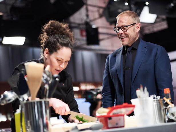 Host Alton Brown watches Chef Stephanie Izard preparing her dish, Duck Tartare, for the Chairman Challenge, as seen on Iron Chef Gauntlet, Season 1.