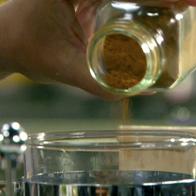 A hand is shaking ground cinnamon out of a glass jar into coffee that is in a French press coffee maker.