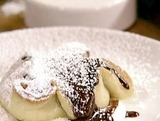 A view of a chocolate pastry that has powdered sugar on top of it. The pastry is sitting on a white plate. The chocolate is in a small light tan pastry that has been cooked. The chocolate is melted and is running over the side of the pastry. The pastry is sitting on some sour cream. The plate is sitting on a light brown wood surface.